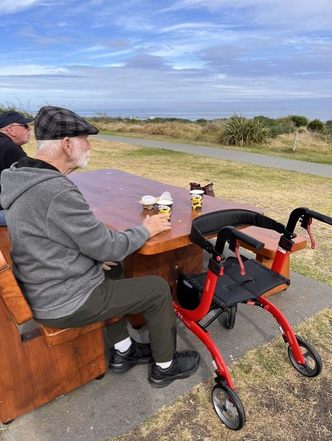 Two older men enjoying refreshments at a seaside picnic table with a mobility walker